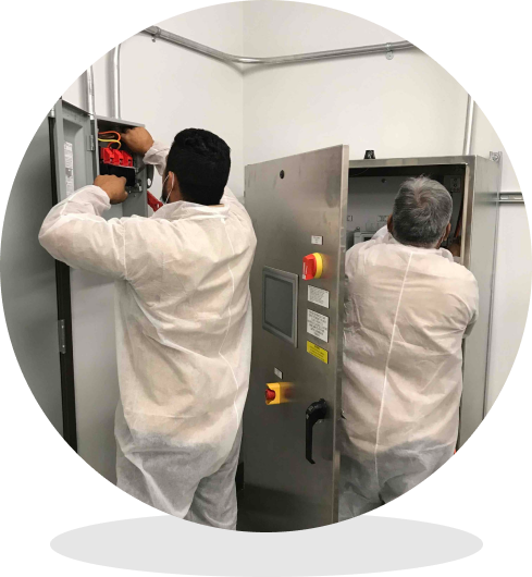 Two technicians in white coveralls service electrical control panels in a utility room.