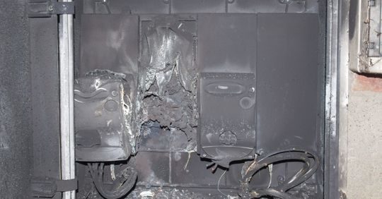 Burnt electrical panel with melted insulation and exposed wires, showing fire damage and soot around the wiring.