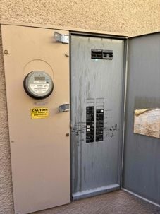 Outdoor electrical meter and service panel in a beige metal enclosure mounted on a wall beside a gray steel door.
