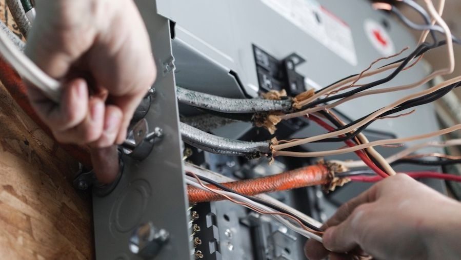 Hands wiring a metal electrical panel with tangled copper cables and insulation.