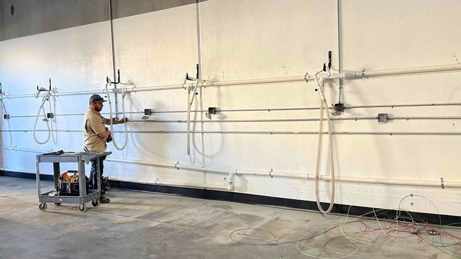 Maintenance worker in a beige shirt and cap stands at a wall with multiple hoses and spray nozzles, next to a wheeled tool cart.