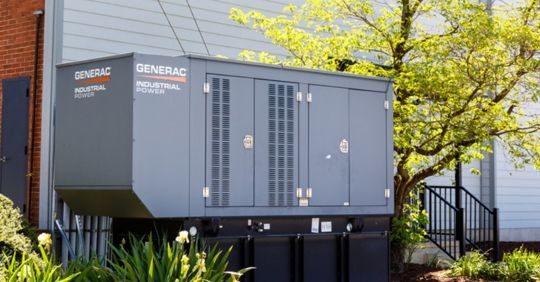An outdoor Generac industrial generator unit mounted on a platform beside a building, with plants in front and a tree nearby.