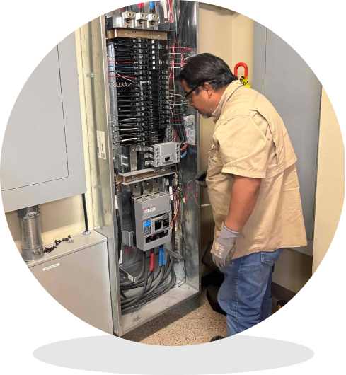 Electrician in gloves and safety glasses inspecting an open electrical control panel with wiring and circuit breakers.