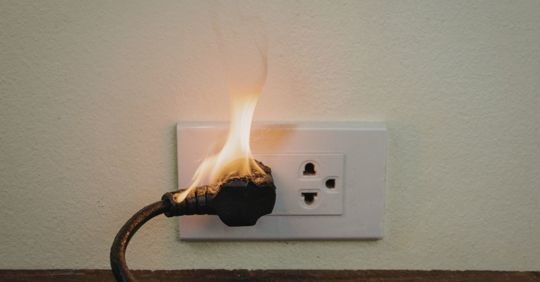 Burned electrical plug in a wall outlet with flames, indicating an electrical fire hazard.