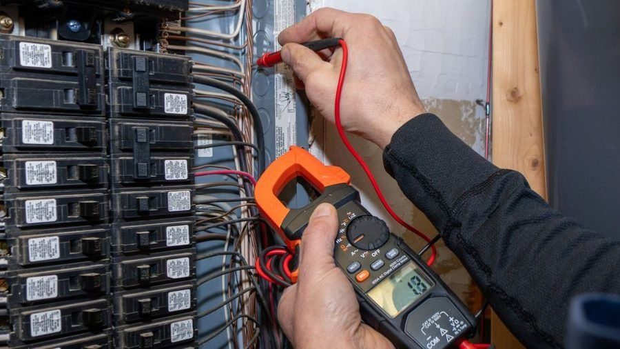 Technician using a digital clamp meter to test wiring inside an electrical breaker panel with red test leads.