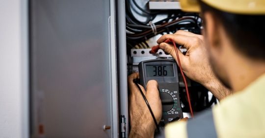 Electrician wearing a hard hat tests an electrical panel inside a metal cabinet using a digital multimeter. Wires are visible and the display shows a reading.