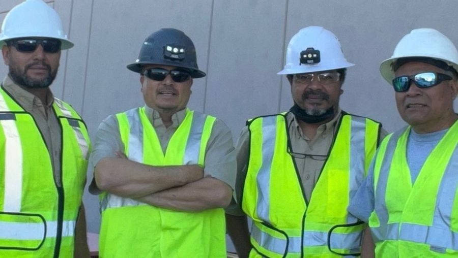 Four construction workers in neon safety vests and hard hats posing for a photo at a work site.