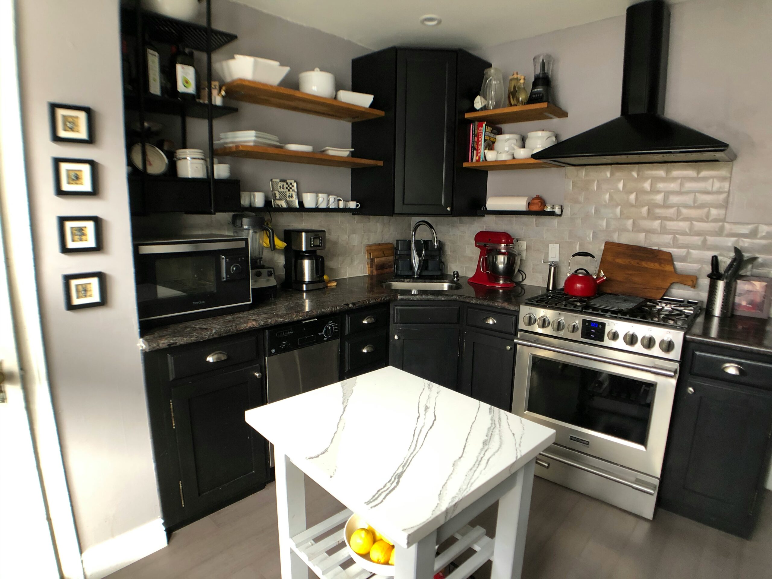 Modern kitchen with black cabinets, open wooden shelves, and stainless steel appliances; white marble island in foreground with lemons stored on a lower shelf.